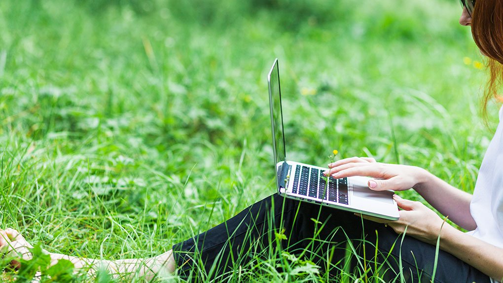 photo représentant une jeune femme assise dans l'herbe et utilisant son ordinateur portable au milieu d'une nature préservée grâce à des sites internets respectueux de l'environnement
